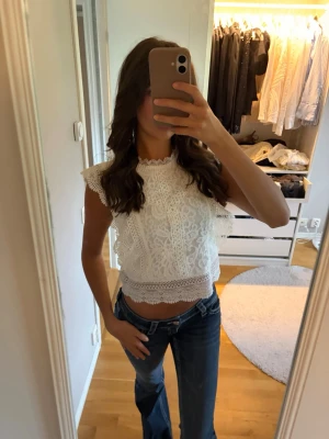 Vit spetsig croppad blus - Säljer en superfin vit croppad blus med spetsdetaljer och volanger längs ärmarna. Toppen har hög hals och är ärmlös, perfekt till jeans eller kjol för en trendig look. Materialet känns lätt och luftigt.