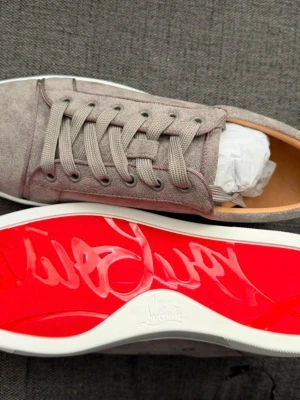 Christian Louboutin 42 - Snygga gråa sneakers i mocka från Christian Louboutin med den ikoniska röda sulan. Skorna har klassisk låg profil, matchande grå skosnören och vita kanter runt sulan. Perfekta för dig som vill sticka ut med stilrena detaljer.