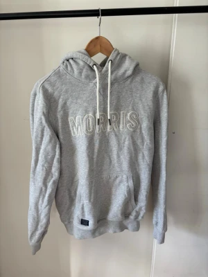 Grå hoodie från Morris - Snygg grå hoodie från Morris med stor vit logga broderad på bröstet. Hoodien har en klassisk känguruficka, ribbade muddar och vit dragsko i huvan. Perfekt för en chill och avslappnad stil.