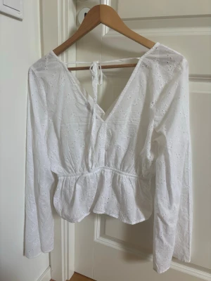 Vit broderad blus med v-ringning - Söt vit blus med broderade detaljer och v-ringning både fram och bak. Blusen har långa ärmar, knytband i nacken och en lätt volang nedtill. Tillverkad i ett tunt, luftigt tyg som passar perfekt till sommaren.
