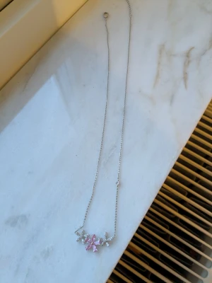 Silverhalsband med rosa blommor - Sött halsband i silverfärg med tunnt kedja och dekorativa blommor framtill. Blommorna är utsmyckade med rosa och klara stenar som ger ett glittrigt intryck. Perfekt accessoar för att lyfta din outfit med en feminin touch.