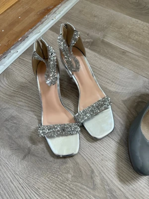 Silvriga pumps med glitterdetaljer - Säljer ett par silvriga pumps med fyrkantig tå och glittriga remmar över foten och runt ankeln. Skorna har öppen häl och en elegant design som verkligen sticker ut. Perfekta för dig som vill ha något extra till outfiten.