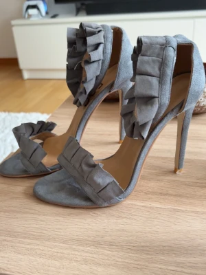 Gråa pumps med volangdetaljer - Säljer ett par gråa pumps med höga smala klackar och snygga volangdetaljer både över tårna och runt ankeln. Skorna har öppen tå och är tillverkade i mockaliknande material. Perfekta för dig som vill sticka ut med en trendig och unik look.
