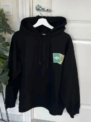 Svart hoodie med spelkortstryck - Säljer en svart hoodie med huva och dragsnören. På bröstet och ryggen finns tryck av två spelkort i grönt och vitt, där ena kortet har texten 'OROLE' och det andra visar en kung. Hoodien har en avslappnad passform och är perfekt för en chill look.