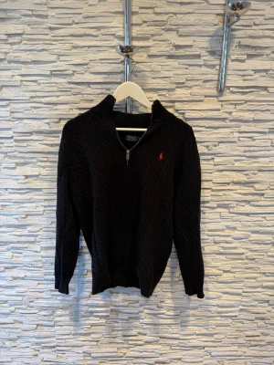 Svart kabelstickad tröja med dragkedja - Svart kabelstickad tröja från Polo Ralph Lauren med halv dragkedja och hög krage. Tröjan har ett rött broderat logomärke på bröstet och ribbade muddar vid ärmslut och nederkant. Perfekt för lager på lager och klassisk stil.