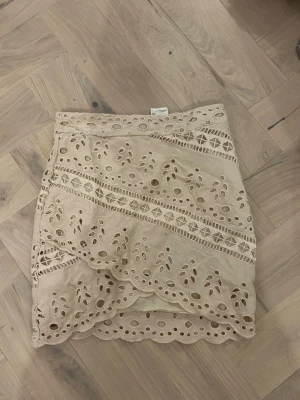 Beige kjol med broderade detaljer - Supersöt beige kortkjol med broderade hålmönster och vågig kant. Kjolen har omlott-design och fina spetsdetaljer som ger en romantisk vibe. Perfekt att styla med en enkel topp för en trendig look.