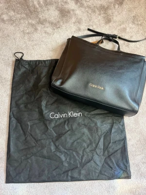 Svart handväska från Calvin Klein - Stilren svart handväska från Calvin Klein i slätt konstläder. Väskan har ett minimalistiskt uttryck med guldfärgad logga framtill, justerbar axelrem och små handtag. Perfekt för dig som gillar klassisk och enkel design.