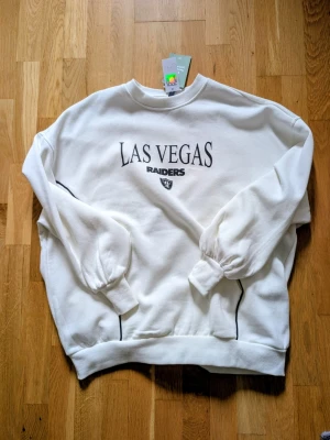 Vit Las Vegas Raiders sweatshirt H&M - Säljer en vit sweatshirt från H&M med trycket 'Las Vegas Raiders' och lagets logga på bröstet. Tröjan har rund hals, ribbade muddar vid ärmslut och nederkant samt svarta detaljer längs sidorna. Perfekt för dig som gillar sportig och avslappnad stil.