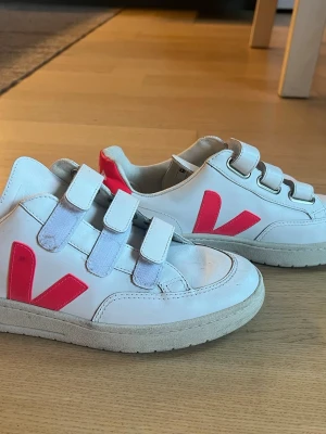 Vita Veja sneakers med kardborreband - Snygga vita sneakers från Veja med tre kardborreband och rosa V-logga på sidan. Skorna har en klassisk låg modell och är tillverkade i läder med gummisula. Perfekta för en clean och trendig look. Använda fåtal gånger.