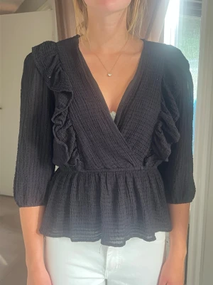Svart omlottopp med volangdetaljer - Snygg svart omlottopp med trekvartsärm och volangdetaljer längs framsidan och axlarna. Toppen har en markerad midja och lätt struktur i tyget som ger en trendig look. Perfekt att styla med jeans eller kjol för en chic outfit. Pris går att diskutera