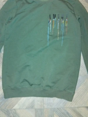 Grön sweatshirt från Amiri - Säljer en grön sweatshirt från Amiri med tryckt logga på bröstet där färgstänk i blått, gult, rosa och svart rinner ner från bokstäverna. Tröjan har rund halsringning och ribbade muddar vid ärmslut och nederkant. Perfekt för dig som gillar streetwear och unika detaljer.