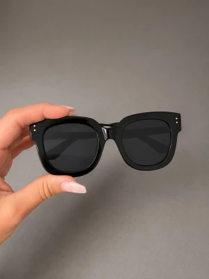 Svarta chunky solglasögon - Säljer ett par chunky svarta solglasögon med fyrkantig form och mörka glas. Bågarna är breda och har små guldfärgade detaljer vid hörnen. Perfekta för en cool och trendig look.