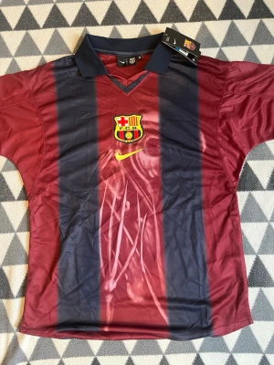 Barcelona matchtröja Nike M - Säljer en officiell FC Barcelona matchtröja från Nike i storlek M. Tröjan är vinröd och marinblå med klubbens emblem på bröstet och Nike-logga i gult. Klassisk krage och V-ringning, perfekt för fotboll eller att visa support.