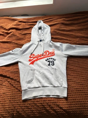 Grå Superdry hoodie med tryck - Säljer en ljusgrå hoodie från Superdry med röd logga och svart tryck på bröstet. Hoodien har stor huva med dragsko, magficka och ribbade muddar vid ärmar och nederkant. Perfekt för en avslappnad och sportig stil.
