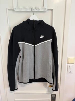 Nike tech fleece svart och grå med dragkedja - Nike hoodie i svart och grått med hel dragkedja framtill och vit logga på bröstet. Hoodien har huva, fickor på sidan och en vit detaljlinje över bröstet. Tillverkad i mjukt sweatshirtmaterial, perfekt för en avslappnad stil.