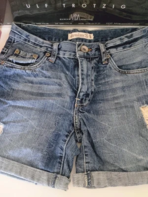Nya Jeansshorts med slitningar denim str 34 Lindex shorts  - Säljer ett par klassiska blå jeansshorts från denim31 med slitna detaljer och upprullade benslut. Shortsen har fem fickor, knappgylf Perfekta för sommaren och ger en cool, avslappnad look. Aldrig kommit till användning så är i fint skick 💖