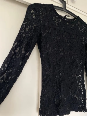 Svart spetsblus med lång ärm - Snygg svart blus i genombruten spets med lång ärm och rund hals. Blusen har ett blommigt spetsmönster som ger en elegant look och är figurnära i modellen. Perfekt att styla med jeans eller kjol för en trendig outfit. Storlek XS