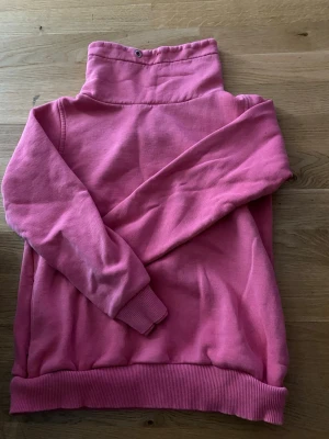 Rosa hoodie med hög krage - En rosa hoodie med hög krage och ribbade muddar vid ärmslut och nederkant. Hoodien har en metallöljettdetalj i kragen och är gjord i mjukt sweatshirtmaterial. Perfekt för en avslappnad och trendig look.