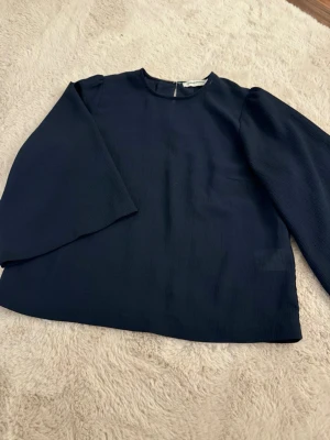 Mörkblå blus - Stilren mörkblå blus från Carin Wester med rund halsringning och långa ärmar. Blusen har en lätt strukturerad yta och stängs med hake i nacken. Perfekt till jeans eller kjol för en clean look.