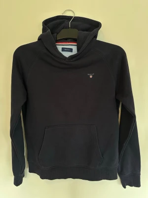 Mörkblå hoodie från Gant - Snygg mörkblå hoodie från Gant med klassisk huva och känguruficka framtill. Liten Gant-logga broderad på bröstet och ribbade muddar vid ärmslut och nederkant. Tillverkad i mjukt bomullsmaterial för skön känsla. Sparsamt använd. 