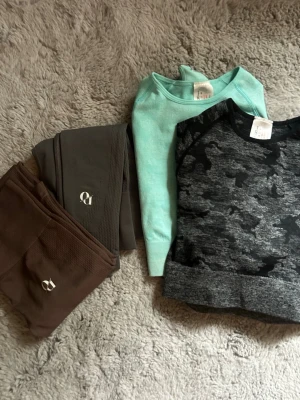 Träningskläder, 2 par tröjor från Boohoo & 2 par tights från OWNU - Två par helt oanvända tights från Hanna Öbergs varumärke OWNU FIT i färgerna mörkgrå och brun. Materialet är stretchigt och andas, perfekt för träning eller chill. Diskret & stilren logga på låret. De bruna tightsen är i stl Small & de gråa tightsen är i stl XS. Båda träningströjorna från Boohoo är i stl small. 150kr för alla 4 plaggen. 