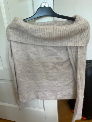 Gina tricot - Mysig beige stickad tröja med bred off shoulder-krage och ribbade detaljer. Tröjan har en avslappnad passform och är perfekt för lager på lager. Materialet ser ut att vara mjukt och skönt, troligen ull- eller kashmirblandning.