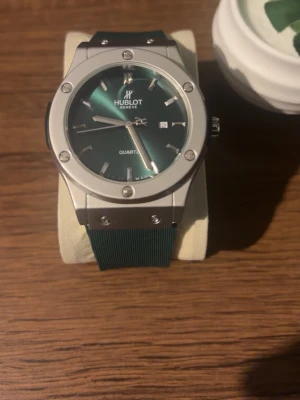 Klocka - Stilren klocka med grön urtavla och silverfärgat boett. Klockan har tydliga index, datumvisning och ett grönt armband i syntetmaterial. Perfekt accessoar för dig som gillar exklusiv och modern design. Priset kan diskuteras!