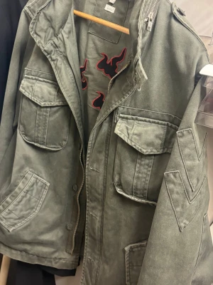 Grön utility/army jacka vintage  - Snygg grön vindjacka i utility-stil med flera stora fickor framtill och dragkedja. Jackan har coola detaljer på ärmen och axlarna, samt en avslappnad passform. Perfekt för dig som gillar streetwear och vill ha en jacka som sticker ut.