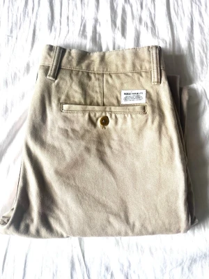 Beige chinos från Norse Projects - Snygga beige chinos från Norse Projects med klassisk rak passform. Byxorna har bälteshällor, sidofickor och en bakficka med knapp. Tillverkade i mjukt bomullstyg som känns skönt mot huden. Perfekta för en clean och stilren look.