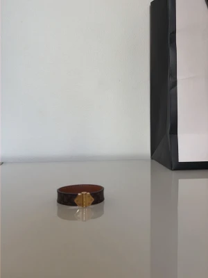 Louis Vuitton brunt läderarmband - Snyggt armband från Louis Vuitton i brunt läder med klassiskt monogrammönster och guldfärgat metallspänne. Armbandet har en elegant och tidlös design som passar perfekt till din vardagsstil. Märkets logga syns på spännet för en exklusiv touch.