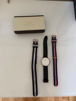 Daniel Wellington klocka med extra band - Stilren klocka från Daniel Wellington med svart läderarmband och två extra tygband i blått/vitt och blått/rosa. Klockan har vit urtavla och guldfärgade detaljer. Perfekt accessoar för en clean look.