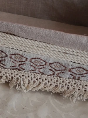 Dekorativ tygkorg med fransar - Säljer en handgjord tygkorg i beige och naturvitt med broderat mönster i brunt. Korgen har dekorativa repdetaljer och fransar längs kanten, perfekt för förvaring av accessoarer eller småprylar. Materialet är textil och bomull.