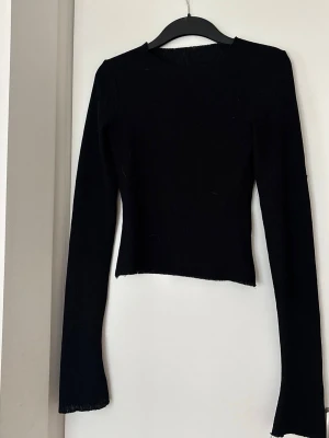 Svart långärmad topp - En svart, ribbad långärmad topp med tight passform och rund halsringning. Toppen har råa kanter vid ärmslut och nederkant, vilket ger en trendig och avslappnad look. Perfekt att styla med jeans eller kjol.