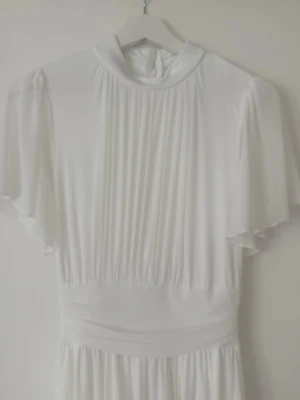 Vit långklänning med plissering - Elegant vit långklänning med plisserad överdel och vida, korta ärmar. Klänningen har hög hals och markerad midja med fina detaljer. Perfekt för dig som vill ha en stilren och klassisk look.