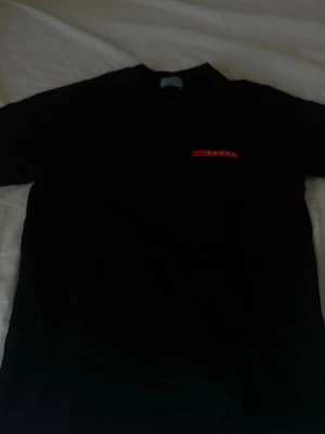 Svart t-shirt från Prada - Snygg svart t-shirt från Prada med klassisk röd logga på bröstet. Enkel och stilren design som passar perfekt till jeans eller shorts. T-shirten har korta ärmar och rund halsringning.