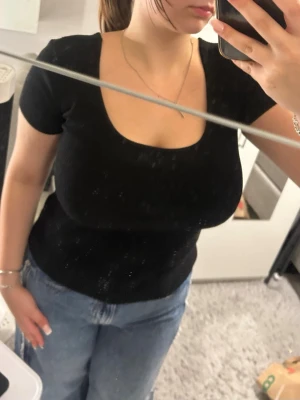 Svart ribbad topp med kort ärm - En svart ribbad topp med korta ärmar och rundad, djup urringning. Snygg och enkel modell som passar till jeans eller kjol. Materialet är stretchigt och sitter tight mot kroppen för en smickrande passform.