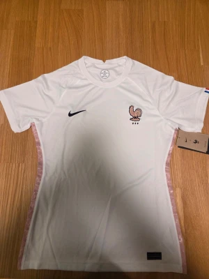 Nike Frankrike slim fit Bortatröja Women's EURO 2022 Dam storlek M - Denna fotbollströja är designad för att ge en bekväm passform. Tröjan har korta ärmar och en rund halsringning. På bröstet finns en broderad logotyp som representerar Frankrikes fotbollsförbund (FFF) tillsammans med Nike-logotypen. En etikett med texten "NOS DIFFÉRENCES NOUS UNISSENT" är placerad i nacken. Ärmsluten har detaljer i rött, vitt och blått, vilket ger en diskret färgklick. Tröjan är utrustad med Nike:s "Engineered"-teknologi, vilket framgår av en etikett längst ner på tröjan. En anna