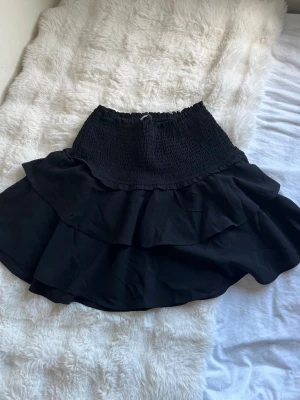 Svart volangkjol med smock - Svart kjol med bred smock i midjan och dubbla volanger som ger en snygg och flowig look. Kjolen är kort och har en lätt, luftig känsla.