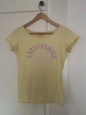 Gul Abercrombie t-shirt med tryck - Gul t-shirt från Abercrombie med rosa text och tryck framtill.
