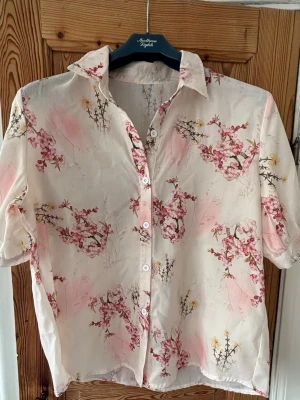 Blommig kortärmad skjorta i siden - Snygg kortärmad skjorta i ljusrosa och vitt med blommigt mönster. Skjortan har klassisk krage, knäppning framtill och är tillverkad i ett tunt, glansigt sidenmaterial. Perfekt för dig som gillar en romantisk och luftig stil.