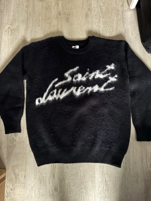 Svart Saint Laurent stickad tröja - Svart stickad tröja från Saint Laurent med vit logotyp i borstigt garn. Aldrig använd. Storlek S men passar även M