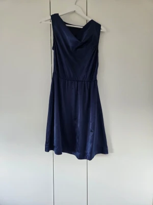 Marinblå ärmlös klänning i satin - Snygg marinblå klänning med v-ringad front och ärmlös design. Klänningen har en lätt glansig satinfinish och en markerad midja som ger en feminin siluett. Perfekt för dig som vill ha en stilren och elegant look.