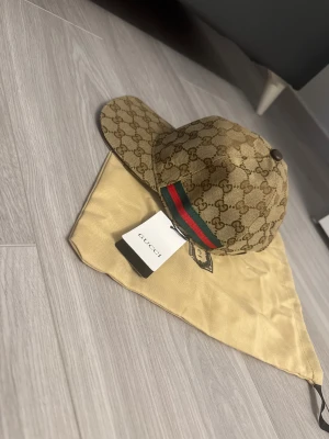 Beige canvas keps  - Snygg keps  i beige med klassiskt monogram över hela kepsen. Har en grön och röd rand på sidan och justerbar rem baktill. Kepsen är tillverkad i canvasmaterial och levereras med  dustbag och tagg.