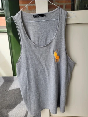 Herrlinne från Polo Ralph Lauren - Snygg grå ärmlös linnetopp från Polo Ralph Lauren med klassisk gul broderad logga på bröstet. Perfekt för varma dagar och enkel att matcha med jeans eller shorts. Mjuk och skön bomullskvalitet.