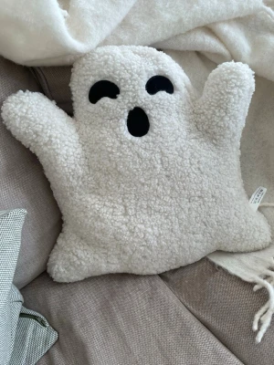 Vit spökformad kudde i teddytyg - Mysig kudde formad som ett spöke i vitt teddytyg med svarta detaljer för ögon och mun. Perfekt som dekoration till halloween eller för att ge rummet en lekfull touch. Kudden är fluffig och har en unik form med utsträckta armar.