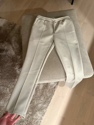 Beige mjukisbyxor Les Deux - Säljer ett par stilrena beige mjukisbyxor från Les Deux med broderad logga och resår i midjan. Byxorna har raka ben, dragsko och mjukt material som känns skönt mot huden. Perfekta för en avslappnad look.