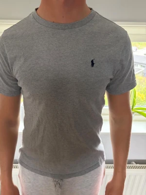 Grå t-shirt från Ralph Lauren - Klassisk grå t-shirt från Ralph Lauren med broderad logga på bröstet. T-shirten har rund halsringning och korta ärmar. Perfekt basic-plagg i mjuk bomull som funkar till allt. Litet hål precis vid märket men inget man tänker på. Barnstorlek L 14-16 men sitter som en vanlig S!!