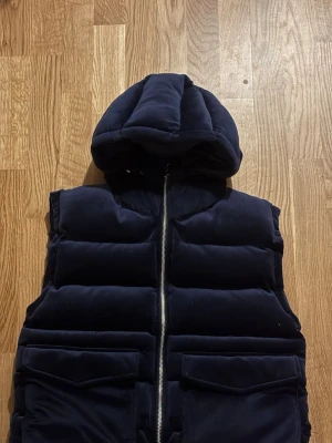 Mörkblå quiltad dunväst med huva - Snygg mörkblå dunväst med quiltad design och stor huva. Västen har två stora fickor framtill och stängs med en silverfärgad dragkedja. Perfekt för lager-på-lager och en cool streetstil.