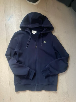 Mörkblå hoodie från Lacoste - Snygg mörkblå hoodie från Lacoste med dragkedja framtill och klassisk krokodil-logga på bröstet. Hoodien har justerbar huva med snören och ribbade muddar vid ärmar och nederkant. Perfekt för en chill och stilren look.