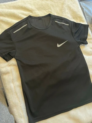 Svart Nike t-shirt med reflexdetaljer - Snygg svart t-shirt från Nike med silverfärgade reflexdetaljer på axlarna och klassisk Nike-logga på bröstet. Tillverkad i ett lätt och funktionellt material som passar perfekt till träning eller en sportig vardagslook. Enkel och stilren design. 173 65 Kg